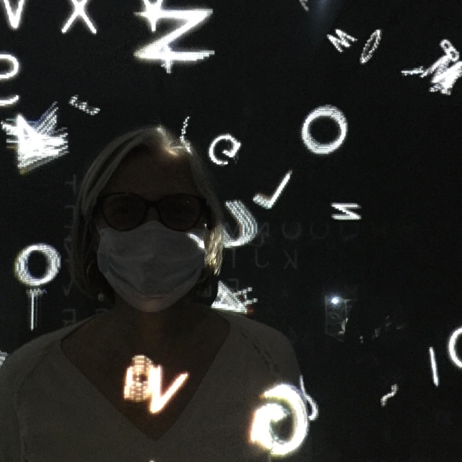 A lady wearing a mask, in the dark, with white letters projected onto her